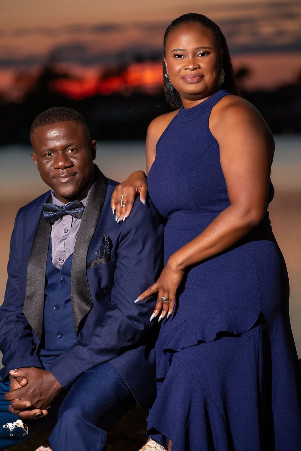A couple in matching navy formal attire poses together during sunset by the water.