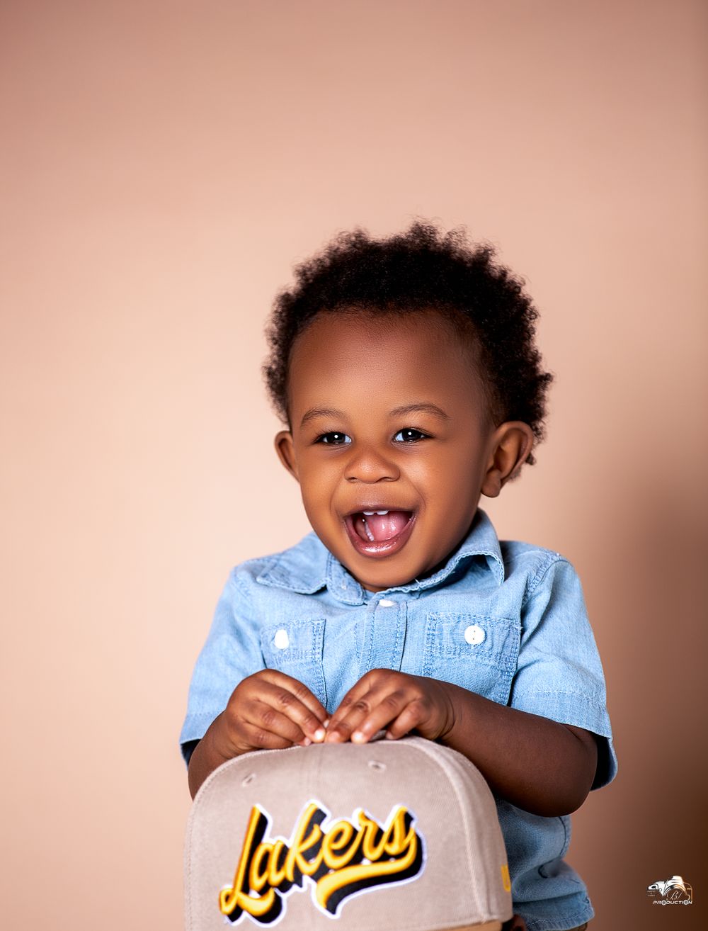 Joyful toddler wearing blue denim shirt holding Lakers merchandise against peach background.