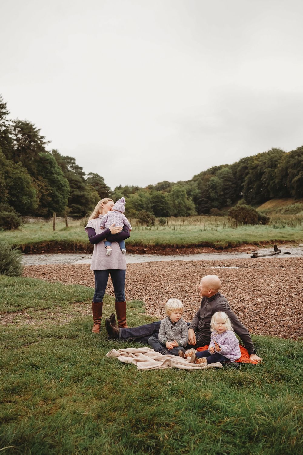 Family enjoying an outdoor picnic by a river, with two toddlers on a blanket and a woman holding a baby.