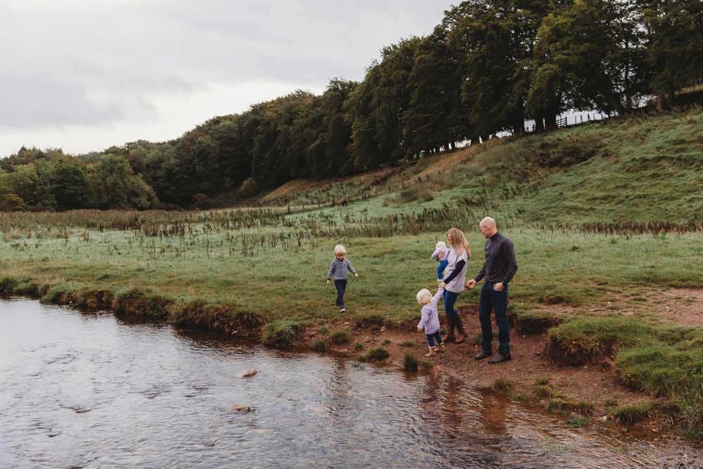 Family of four standing by a peaceful river in a lush green countryside landscape on a cloudy day.
