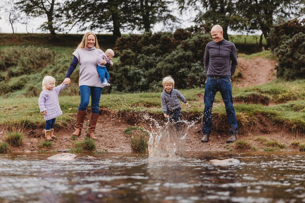 Family of five enjoying a splash by a stream outdoors, with children playing in water while parents watch and smile.