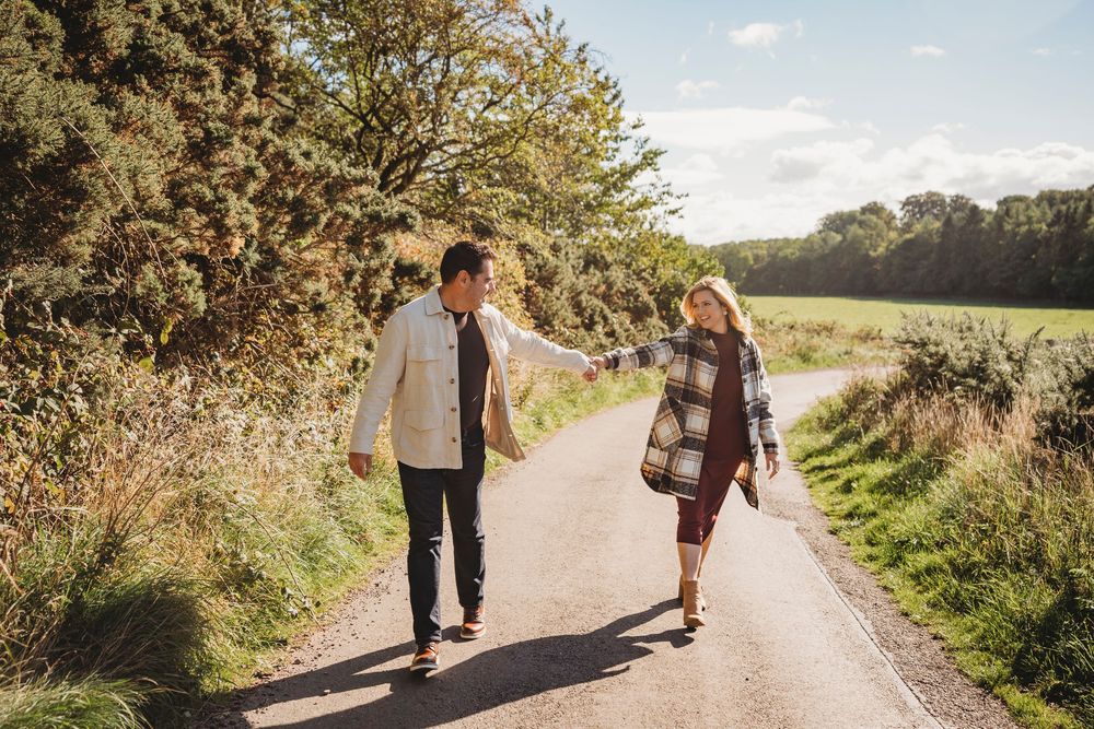 Couple walking hand in hand on a scenic country road surrounded by lush green trees and fields on a sunny day.