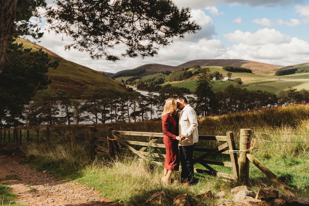 Couple sharing a romantic moment by a wooden gate with scenic Scottish hills and a loch in the background.