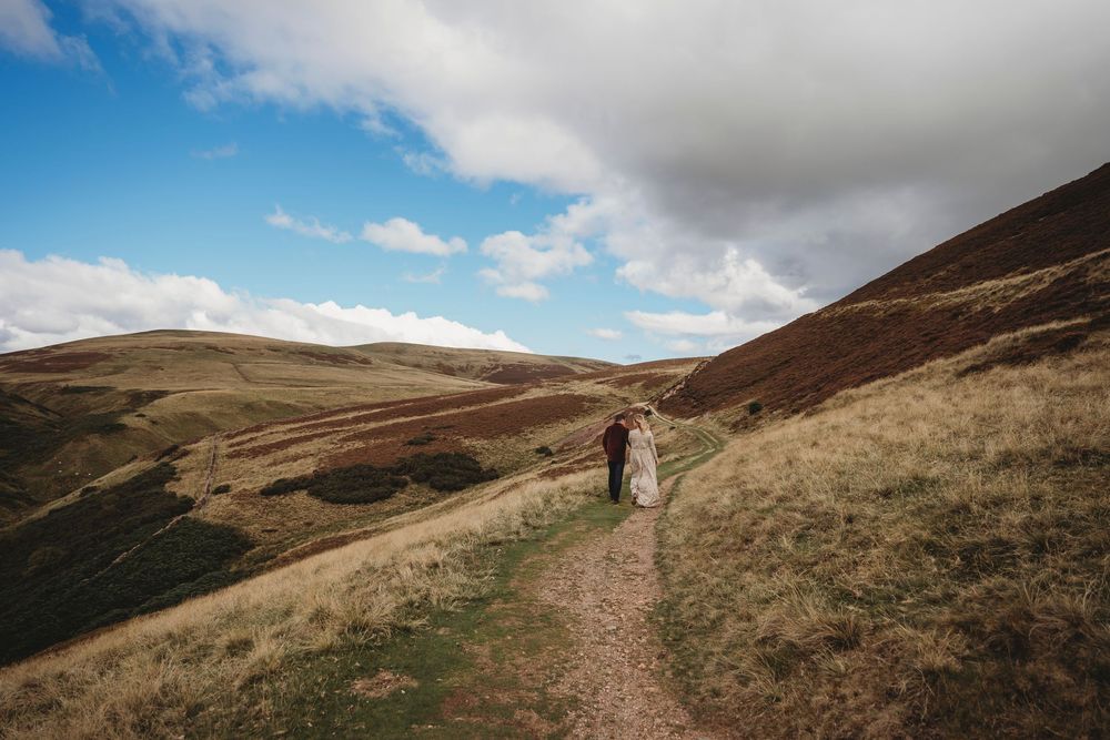 A couple walks a scenic highland trail under a blue sky, surrounded by rolling hills and golden moorland.