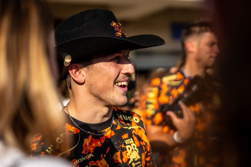 Smiling young man in black cowboy hat and orange floral shirt at a motorsport event.