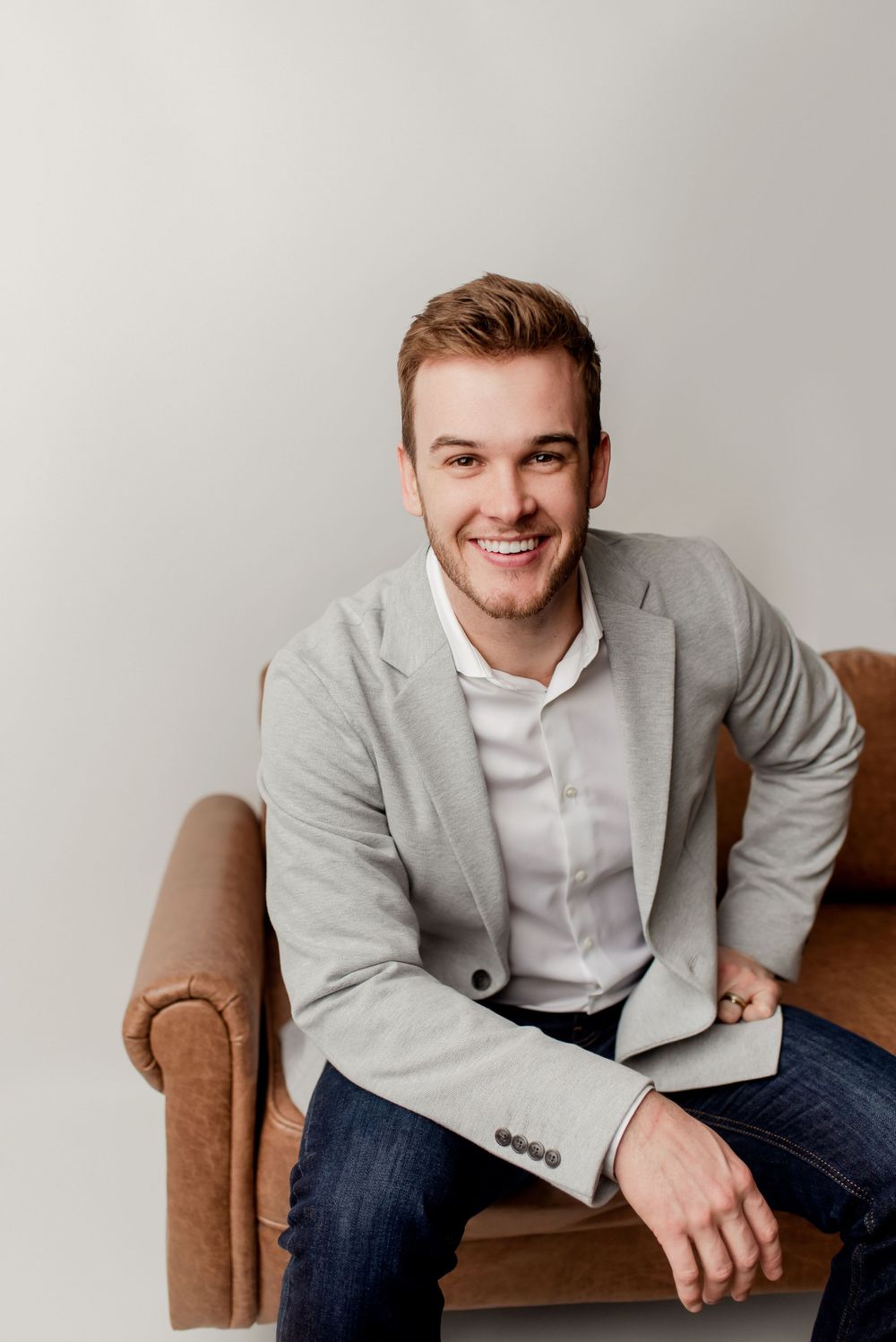 Smiling young man in gray blazer and jeans sitting relaxed on leather chair against white background.