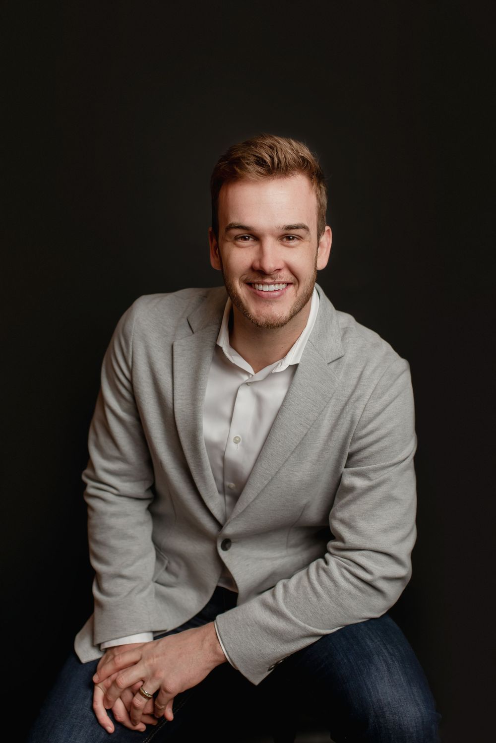 Confident young man in gray blazer smiling warmly against dark studio background.