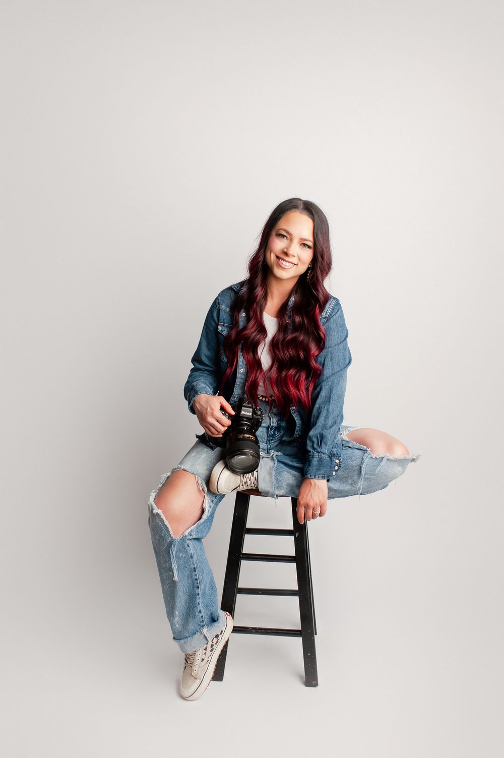 A smiling female photographer sits on a stool holding a camera wearing ripped jeans and a denim jacket.