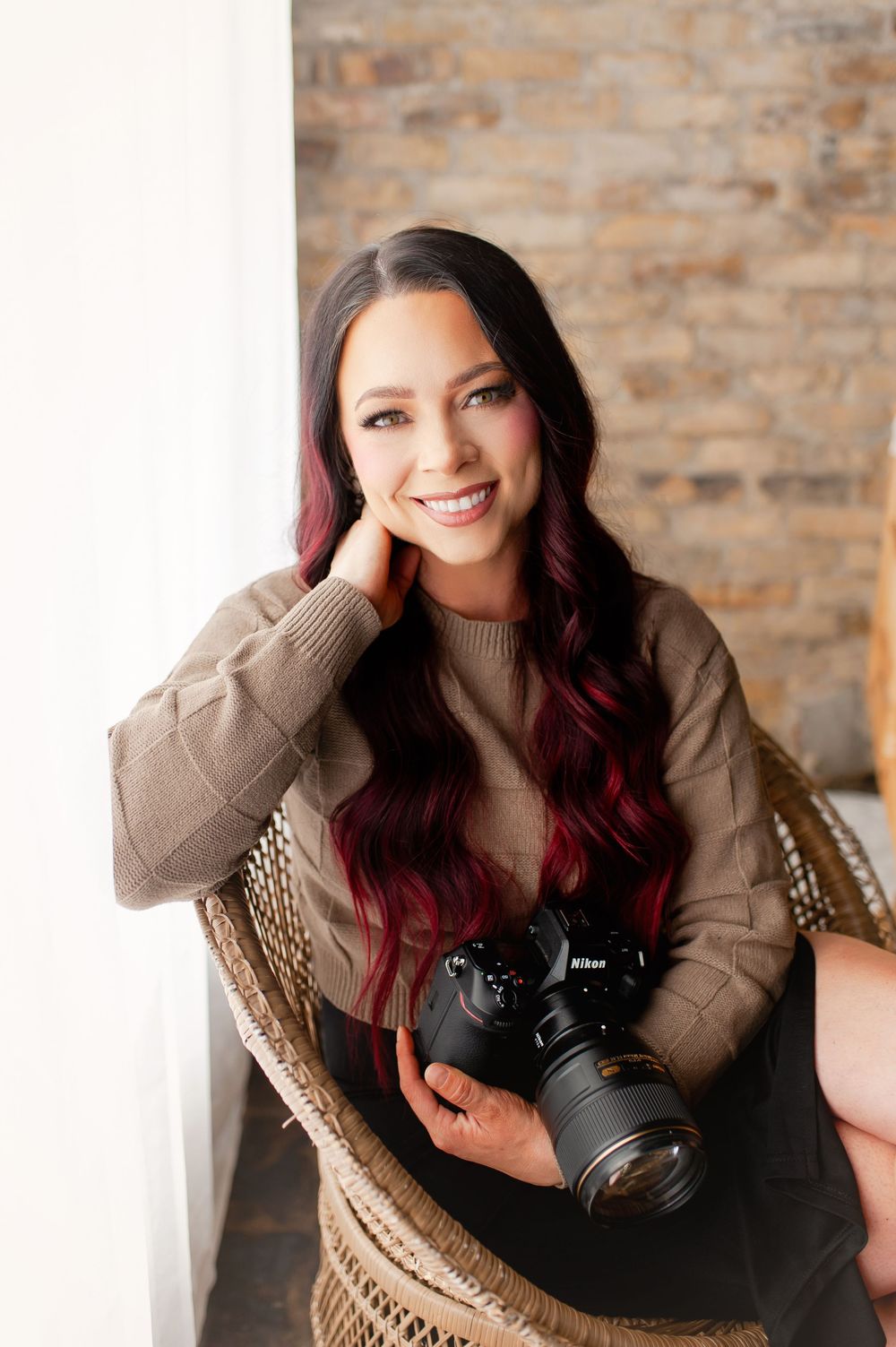 A smiling female photographer with red-highlighted hair holds a Nikon camera while seated in a wicker chair.
