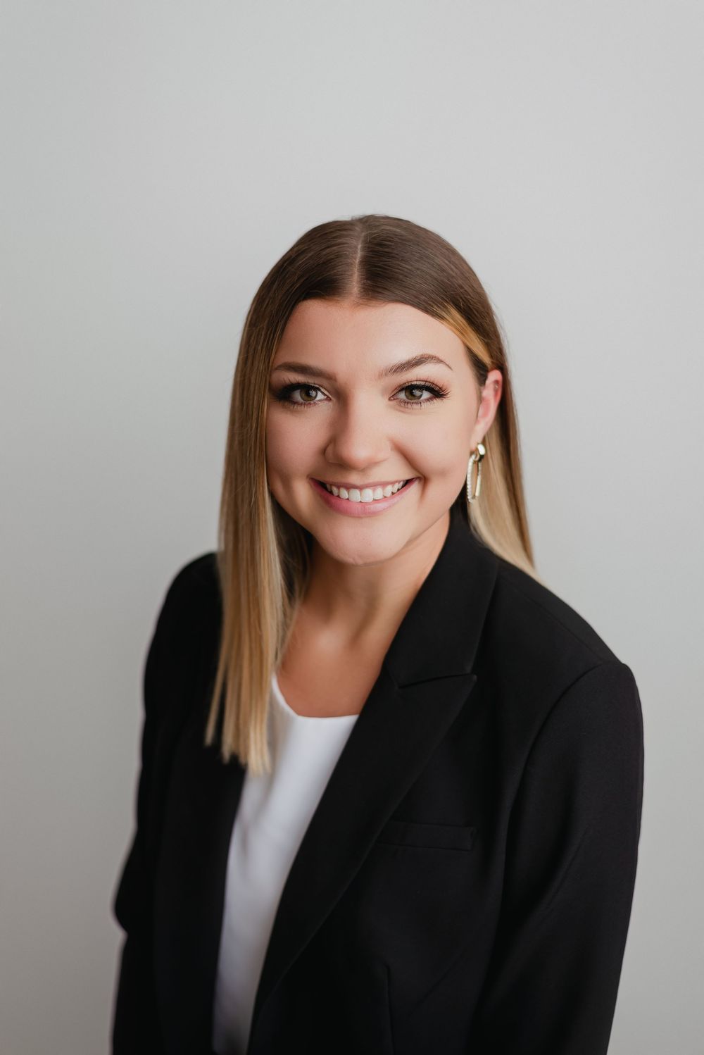 Professional headshot of smiling blonde woman in black blazer against light gray studio background.