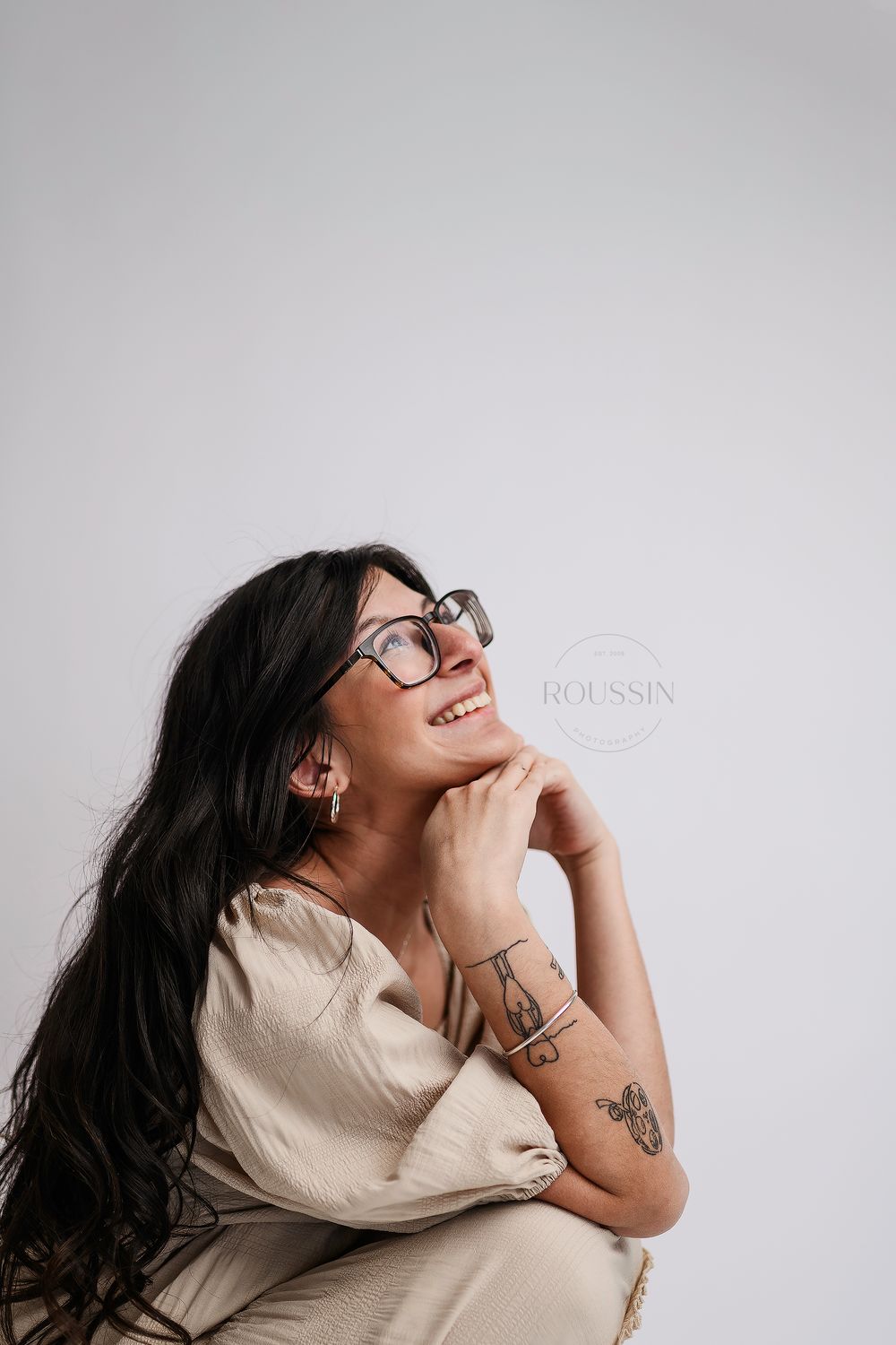 Smiling woman with glasses and tattoos in beige outfit posing against light gray background.