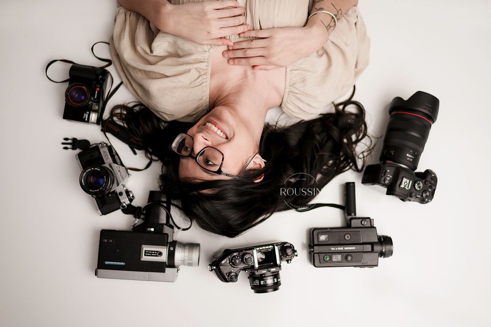 Female photographer lying on floor surrounded by various cameras and photography equipment.