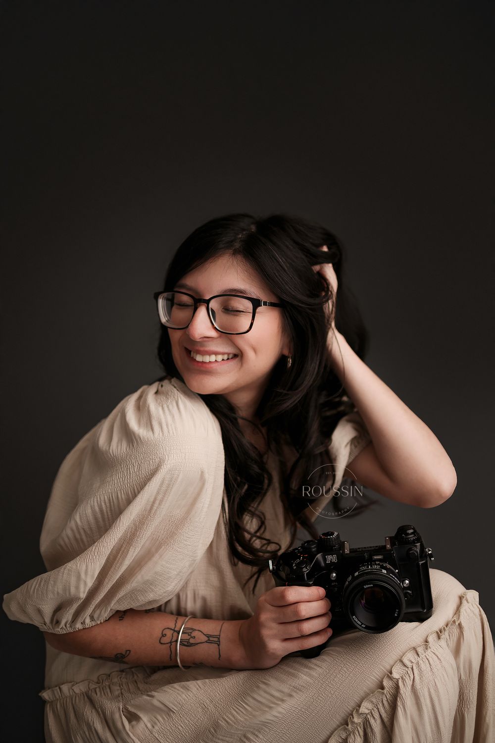 Smiling female photographer with glasses holding camera against dark studio background.
