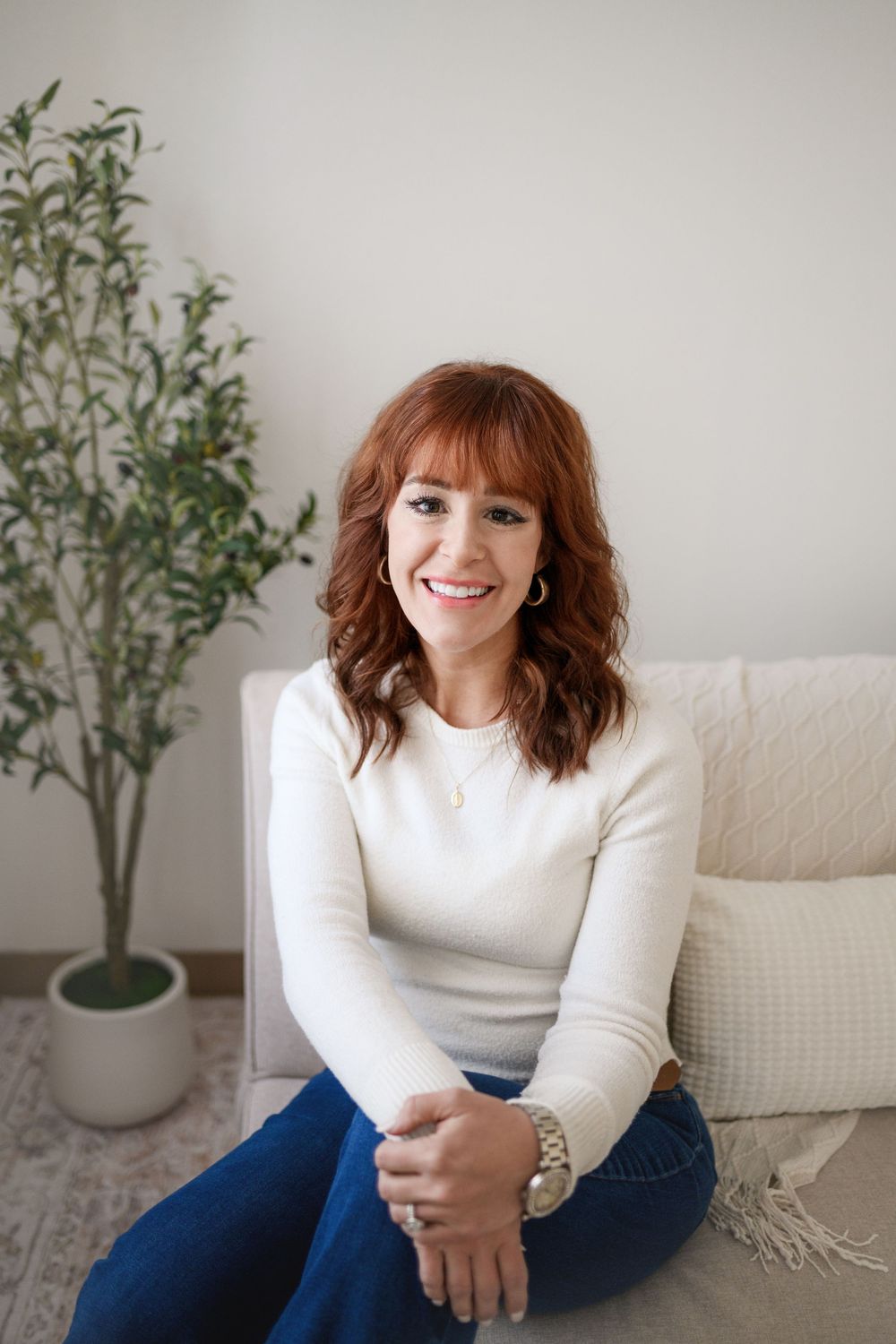 Smiling redhead woman in white sweater and jeans sitting on floor near couch.