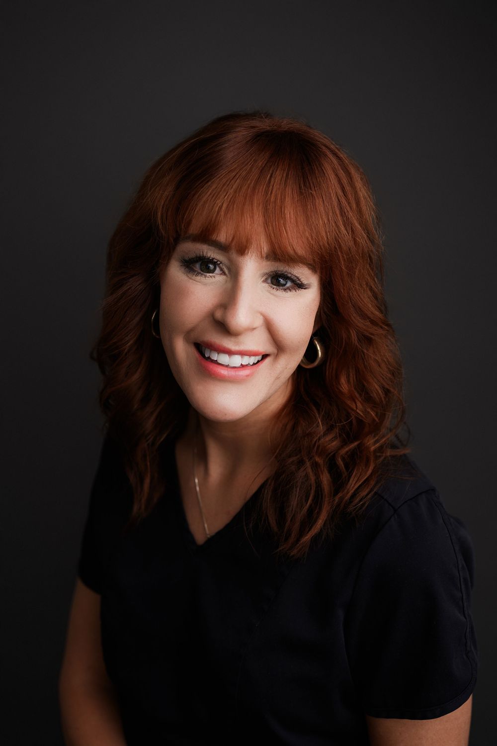 Professional headshot of smiling redhead woman in black top against dark background.
