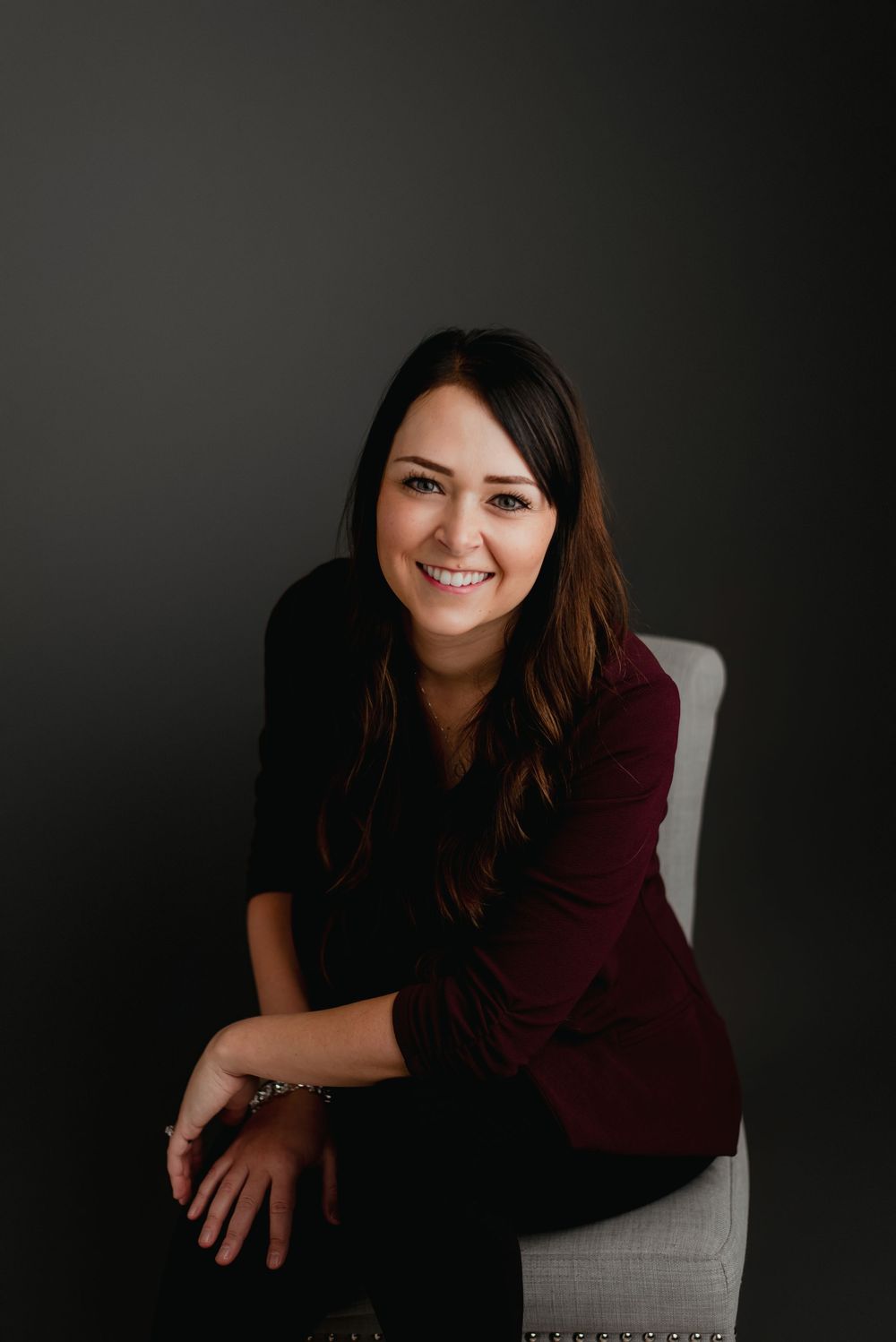 Professional headshot of smiling brunette woman in maroon top seated in chair against dark background.