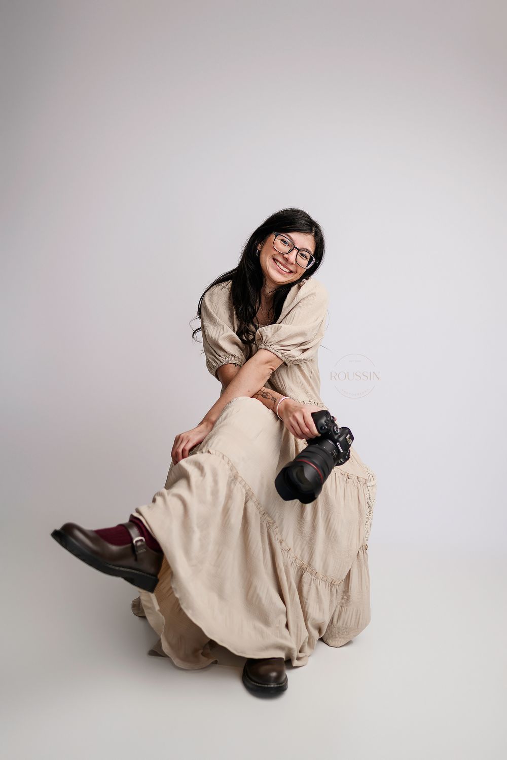 Smiling female photographer in beige dress sitting on floor holding professional camera.