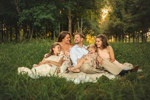 A family relaxes together on a white blanket in tall grass during golden hour sunset in a wooded area.