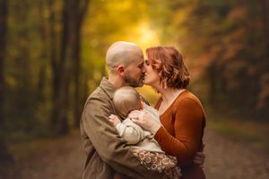 Mom and dad kiss while holding toddler outdoors during a fall photo session with warm golden lighting by Marietta photographer.