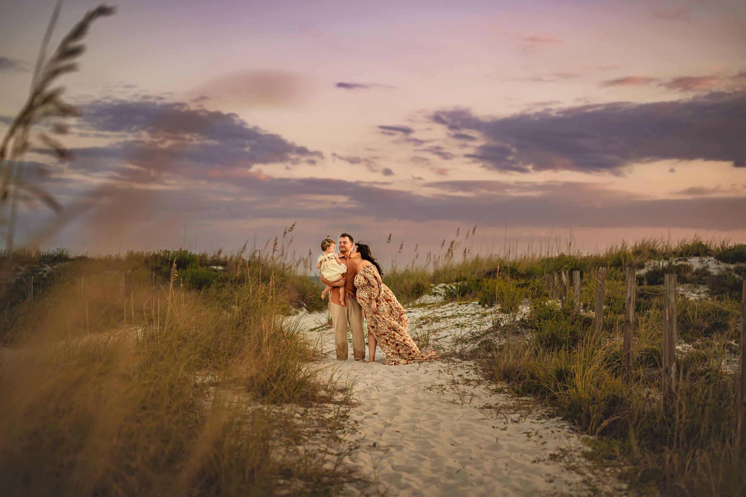 A pregnant mom and dad embraces toddler on a sandy beach path at sunset with sea oats swaying in the foreground and  purple sky.