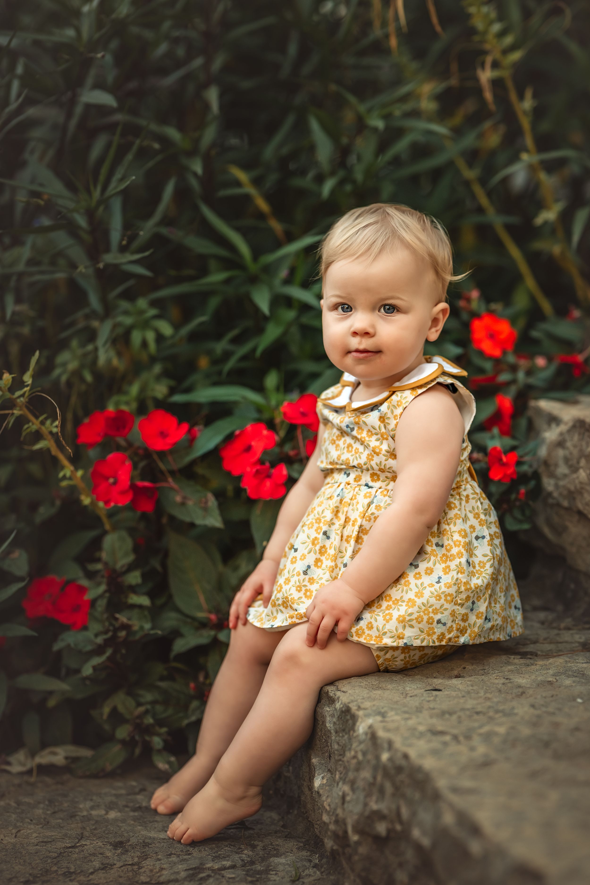 A baby in a yellow floral dress sits on stone stairs with red flowers blooming in the background.