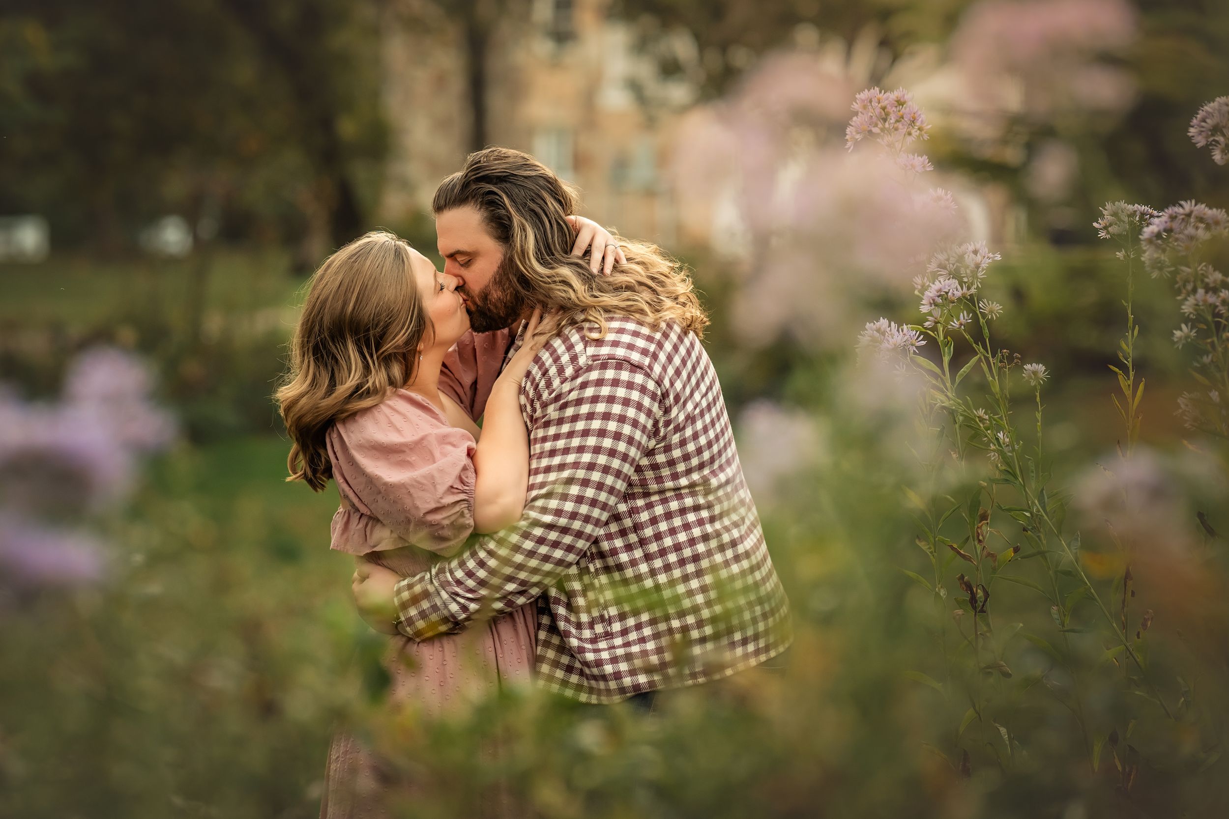 Couple embraces in a romantic outdoor setting framed by purple flowers and greenery with golden sunlight filtering through trees.