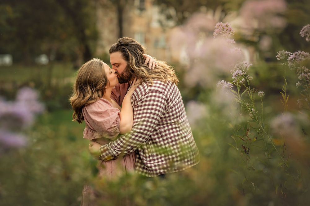Couple embraces in a romantic outdoor setting framed by purple flowers and greenery with golden sunlight filtering through trees.