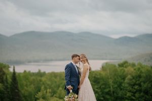 A couple in wedding attire stands on a hilltop overlooking a misty mountain lake surrounded by evergreen forest.