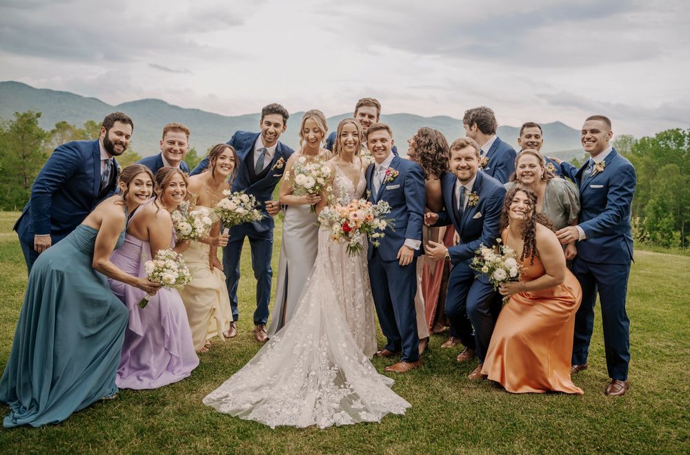 Wedding party in navy suits and colorful bridesmaid dresses pose together on a grassy hillside with mountains in background.