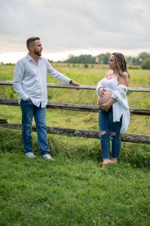 A maternity photoshoot in a rustic setting with a wooden fence and green meadow backdrop.