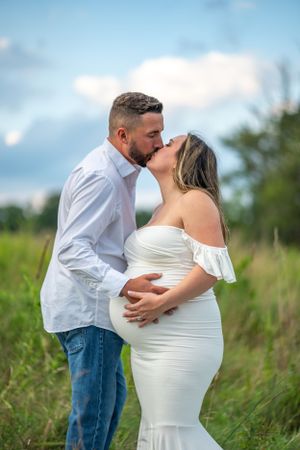 Intimate maternity portrait of a couple in white attire standing in a sunlit field.