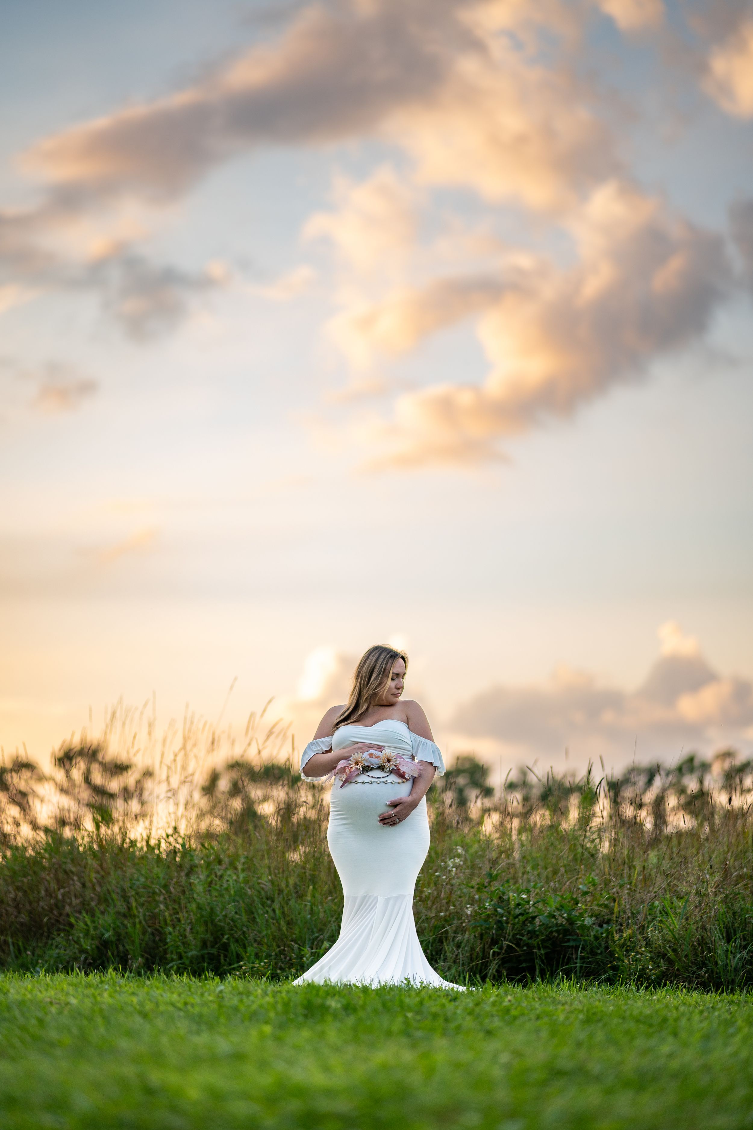 Elegant maternity portrait of woman in white dress standing in field during golden hour.