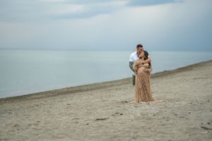 A romantic couple embraces on a serene beach at dusk with calm water and cloudy sky in the background.