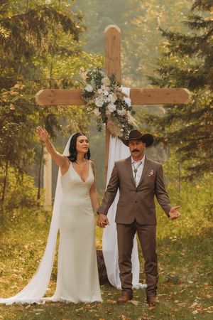 A couple celebrates in front of a wooden cross decorated with white flowers in a sunlit forest.