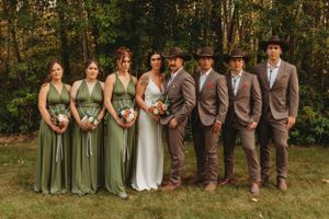 Wedding party in sage green dresses and gray suits poses together in woodland setting.