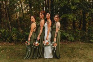 Bridesmaids in sage green dresses pose with bride in white gown against forest backdrop.