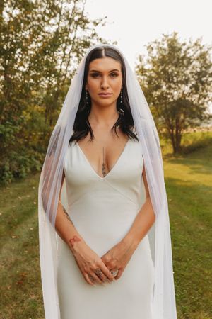 Bride in sleek white wedding dress and veil poses in outdoor garden setting.