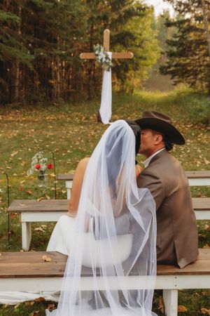Couple sits on wooden bench near decorated cross during outdoor wedding ceremony.