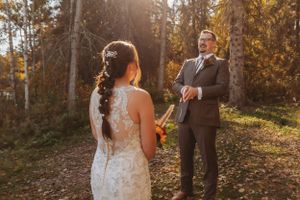A couple stands together during their autumn wedding ceremony surrounded by sunlit trees.