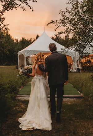A couple walking towards a white wedding tent at sunset through a garden pathway.