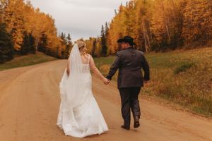 Newlyweds walk hand in hand down a country road surrounded by autumn trees.