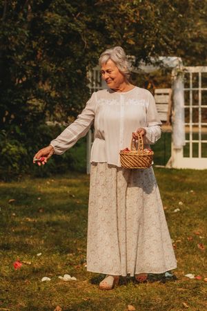 Woman in elegant cream outfit holds a basket while scattering items during an outdoor celebration.