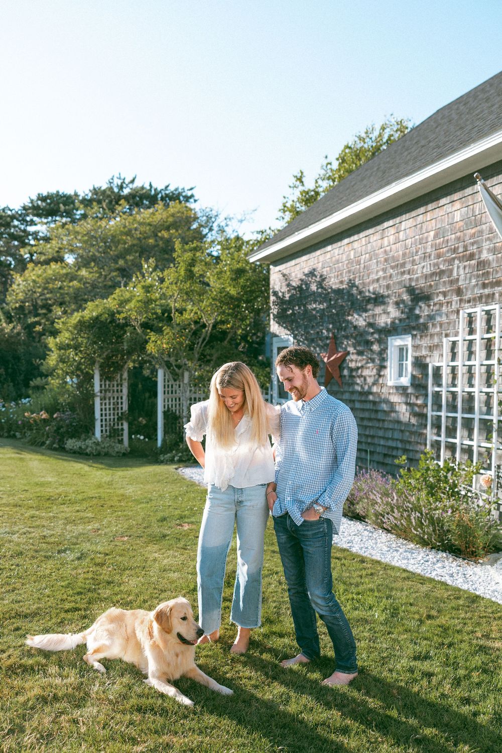 Couple standing in their backyard with their golden retriever beside a shingled house.