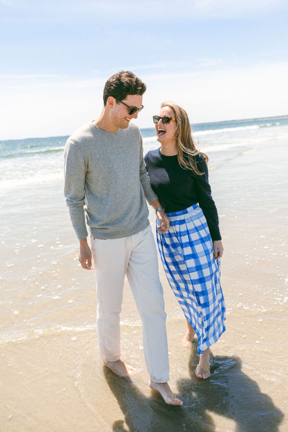 Couple walking barefoot on a sunny beach, man in gray sweater and white pants, woman in black top and blue gingham skirt.