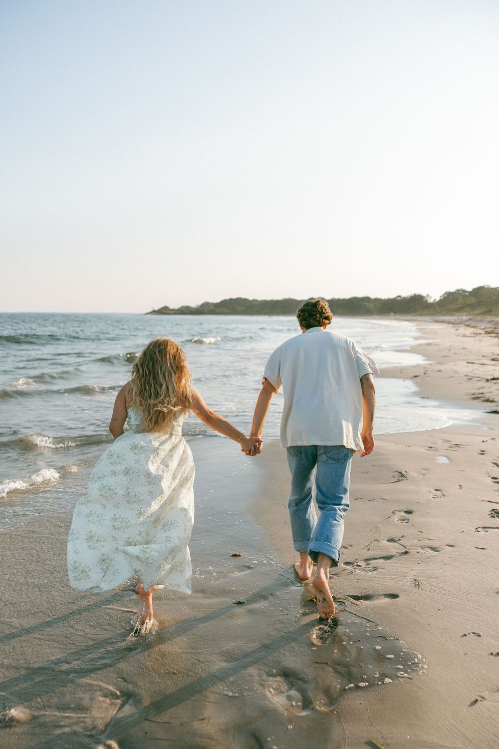 Couple holding hands walking barefoot along sandy beach shoreline in white dress and jeans at sunset.
