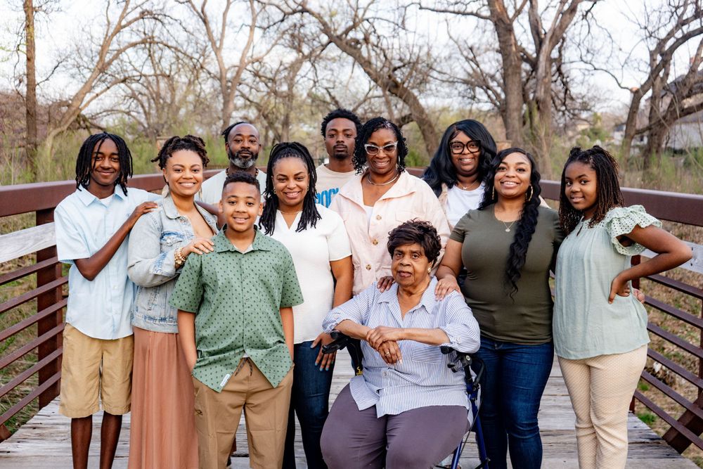 A large multigenerational Black family poses together on a wooden bridge surrounded by bare winter trees.
