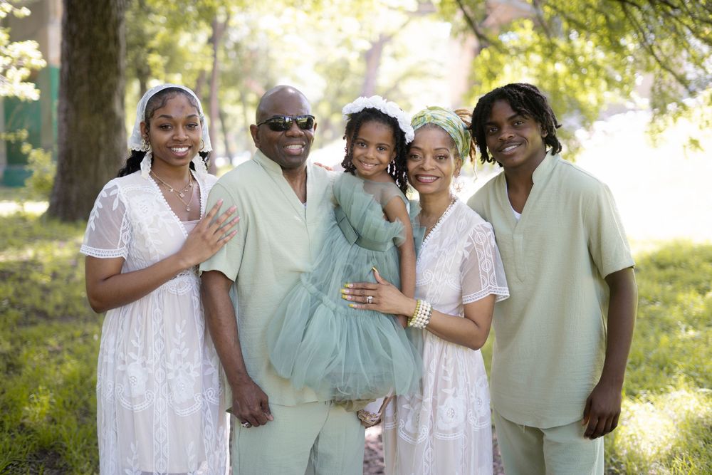 Family of five in mint and white outfits posing together outdoors under green trees in summer.