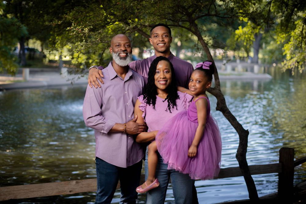 A joyful Black family of four poses near a pond, wearing coordinating purple outfits among lush green trees.