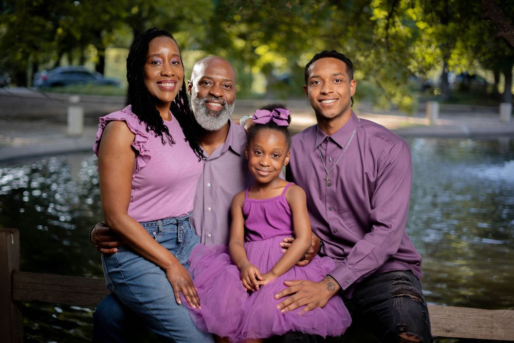 A smiling Black family of four in matching purple outfits sits together near a serene pond outdoors.
