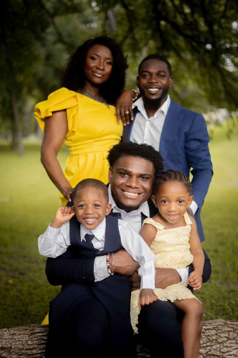 Happy family of five with two parents and three children posing outdoors under green trees in coordinated outfits.